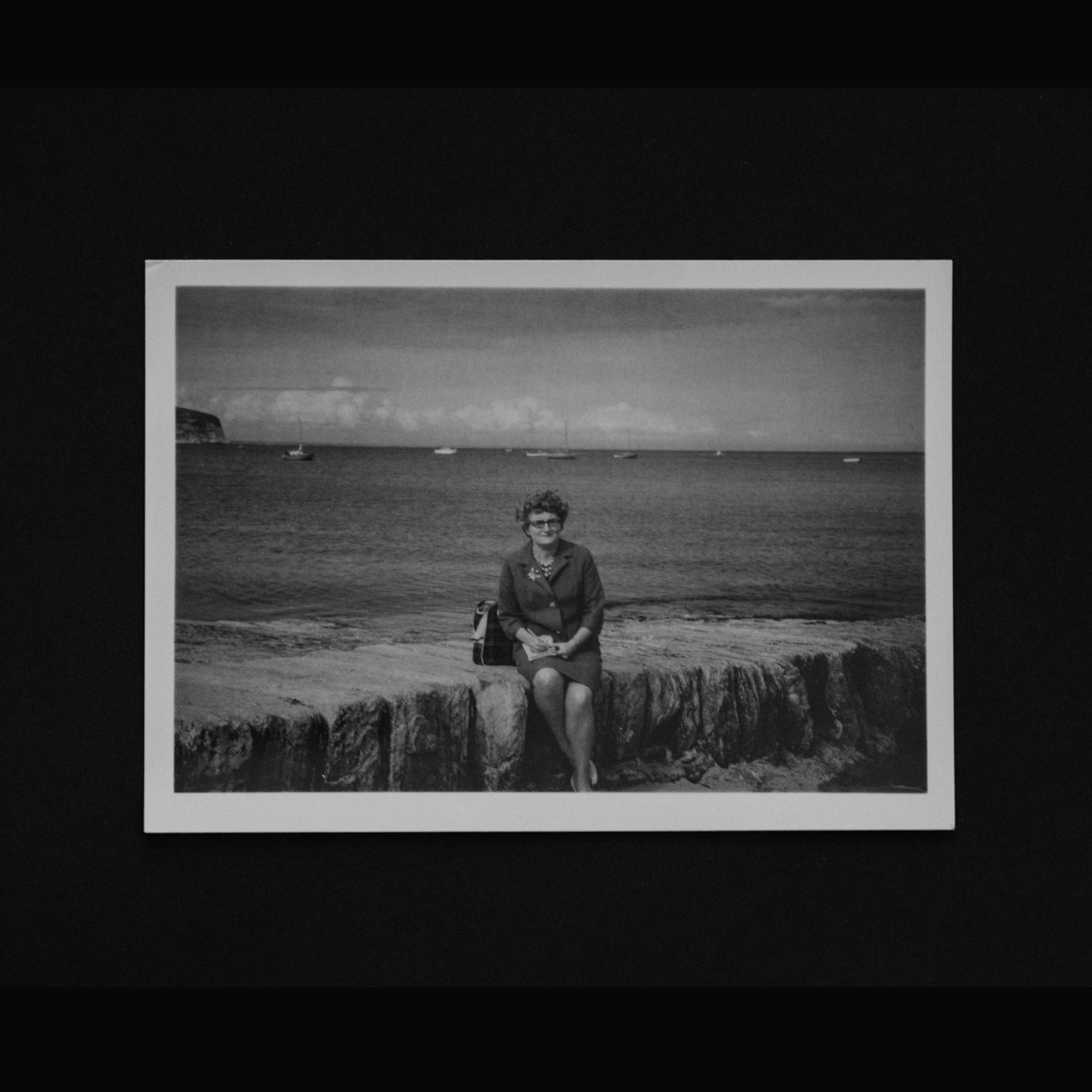 Black and white photo of a woman in a dress and sunglasses sitting on a stone wall by the sea. Boats are visible on the water under a cloudy sky.