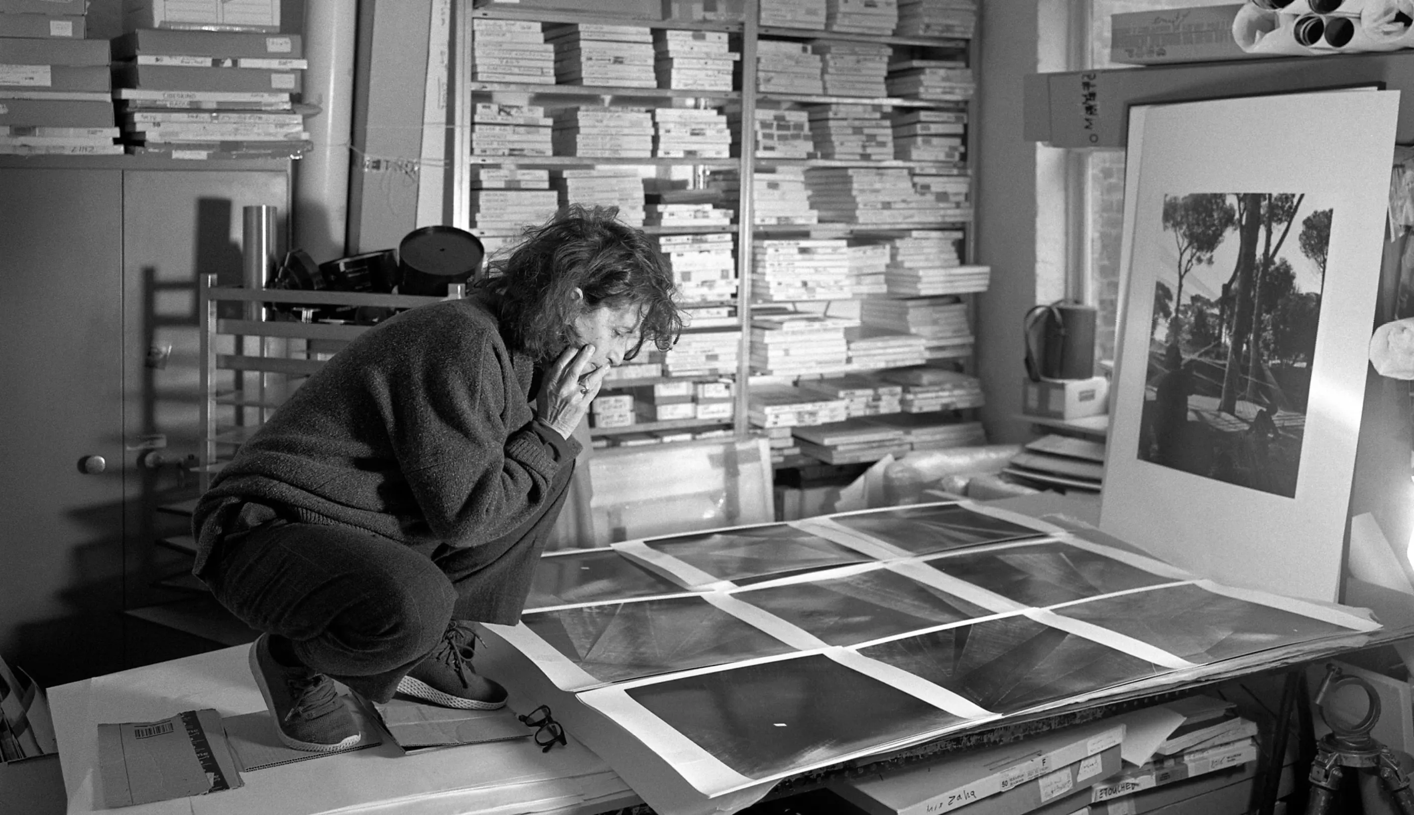 Black and white photo of a woman crouching on a desk, intently looking at several large photographic prints spread before her. Behind her, floor-to-ceiling shelves are packed with boxes and archives in a cluttered studio space.