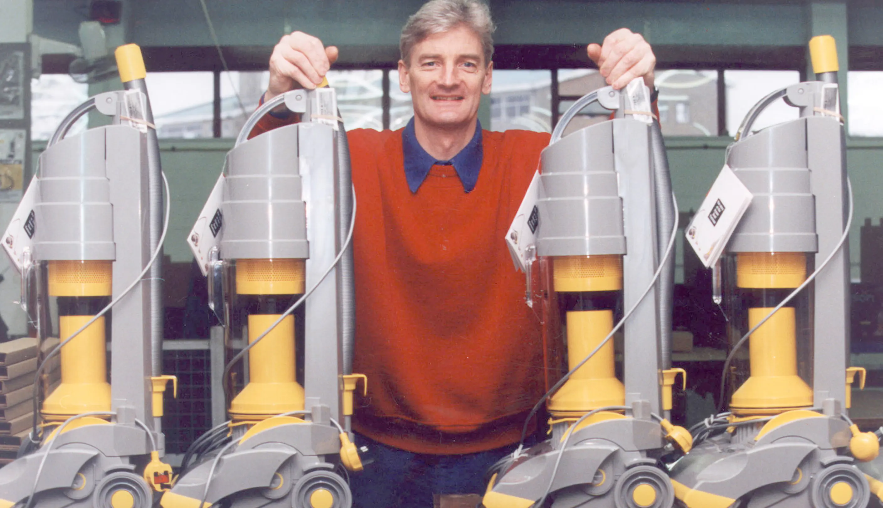 A man in a red sweater and blue collared shirt stands behind four identical grey and yellow upright vacuum cleaners lined up in a row. He has his hands placed on the handles of the middle two vacuums.