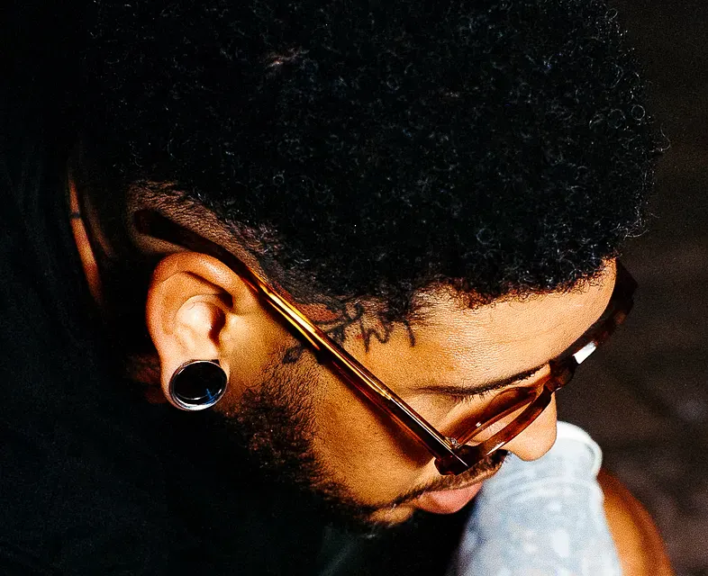 High-angle, close-up profile of a man with short, dark curly hair and a beard, wearing brown-rimmed glasses and a large black ear gauge in his lobe.