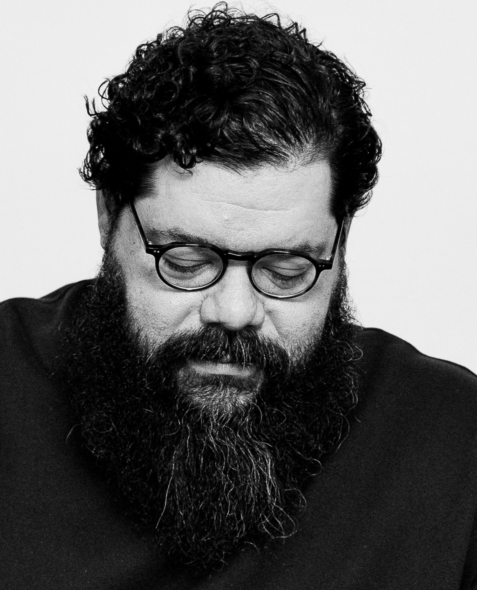 Black and white, close-up portrait of a man looking downward with eyes closed. He has short, curly dark hair, round black-rimmed glasses, a full, thick beard and mustache, and is wearing a dark t-shirt against a plain white background.