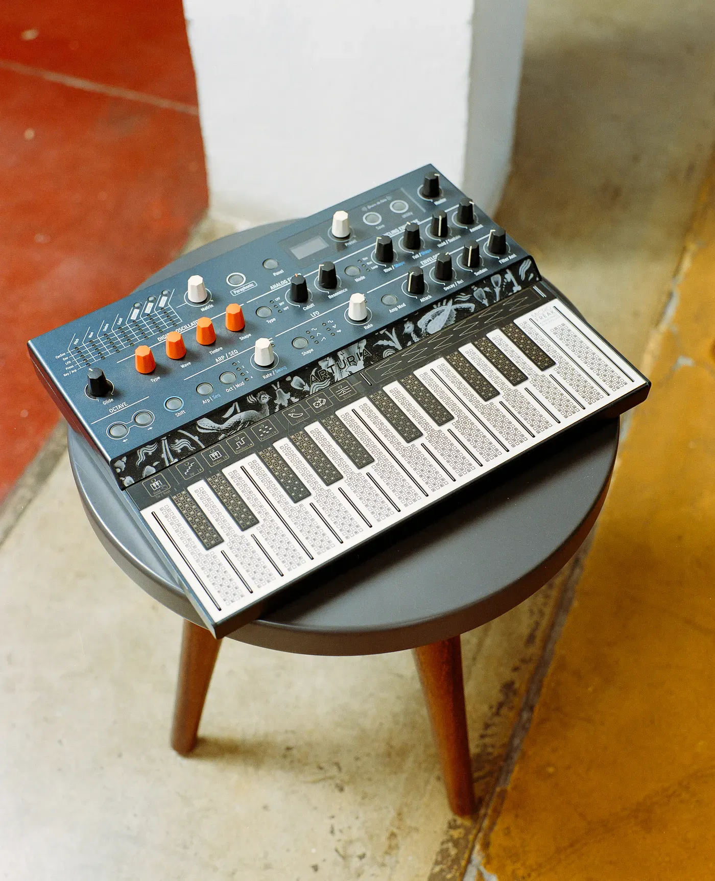 An Arturia MicroFreak synthesizer with its distinctive touch-plate keyboard rests on a small, round grey stool with wooden legs, angled between a red floor and a concrete floor.