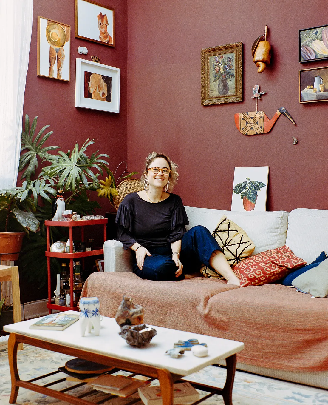 A woman with glasses sits on a couch in a room with dark red walls filled with artwork. In the foreground, a marble coffee table holds small ceramic sculptures, next to a large potted plant and a red rolling cart.
