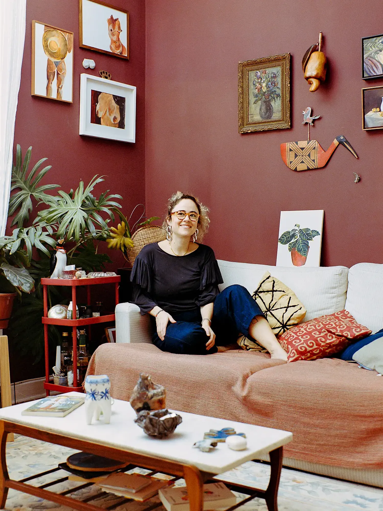 A woman with glasses sits on a couch in a room with dark red walls filled with artwork. In the foreground, a marble coffee table holds small ceramic sculptures, next to a large potted plant and a red rolling cart.