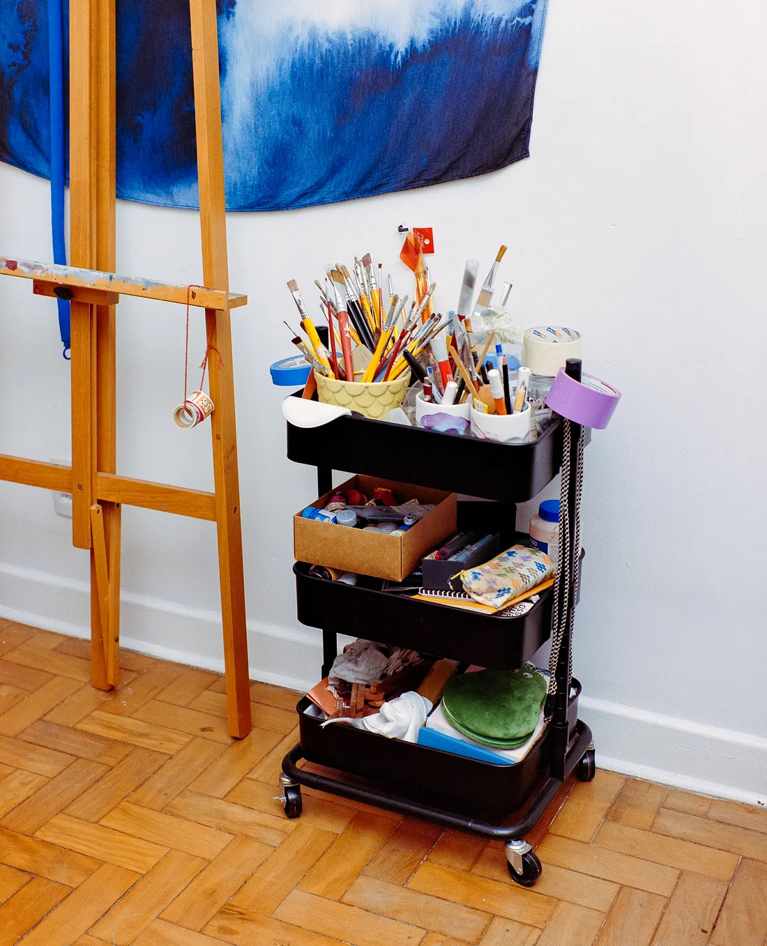 A three-tier black rolling utility cart filled with art supplies stands next to a wooden easel on a parquet floor. The top shelf holds numerous paintbrushes and markers in cups, while the lower shelves contain boxes and various tools.