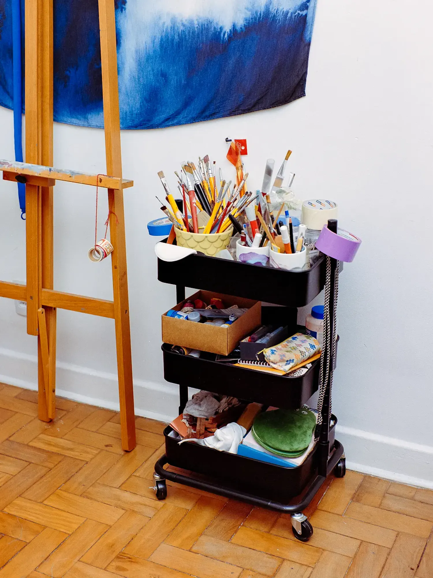 A three-tier black rolling utility cart filled with art supplies stands next to a wooden easel on a parquet floor. The top shelf holds numerous paintbrushes and markers in cups, while the lower shelves contain boxes and various tools.