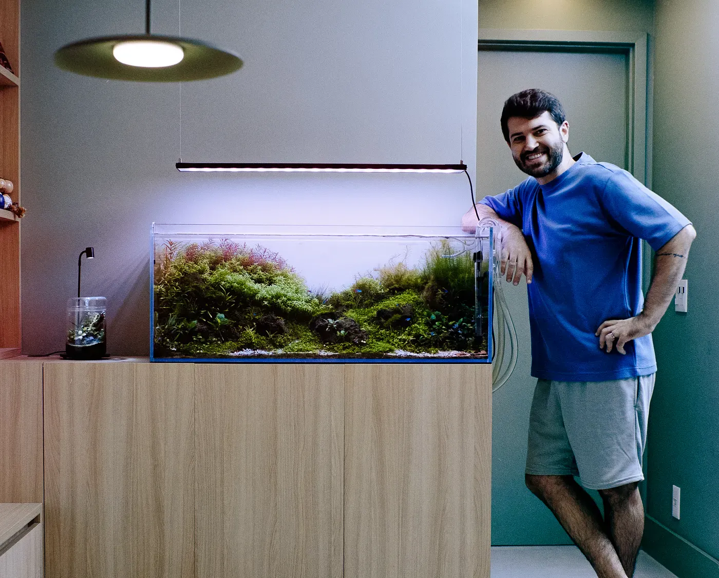 A smiling man in a blue t-shirt and grey shorts leans against a wooden cabinet supporting a large, brightly lit rimless aquarium filled with lush green aquatic plants. A sleek LED light fixture hangs directly above the tank.