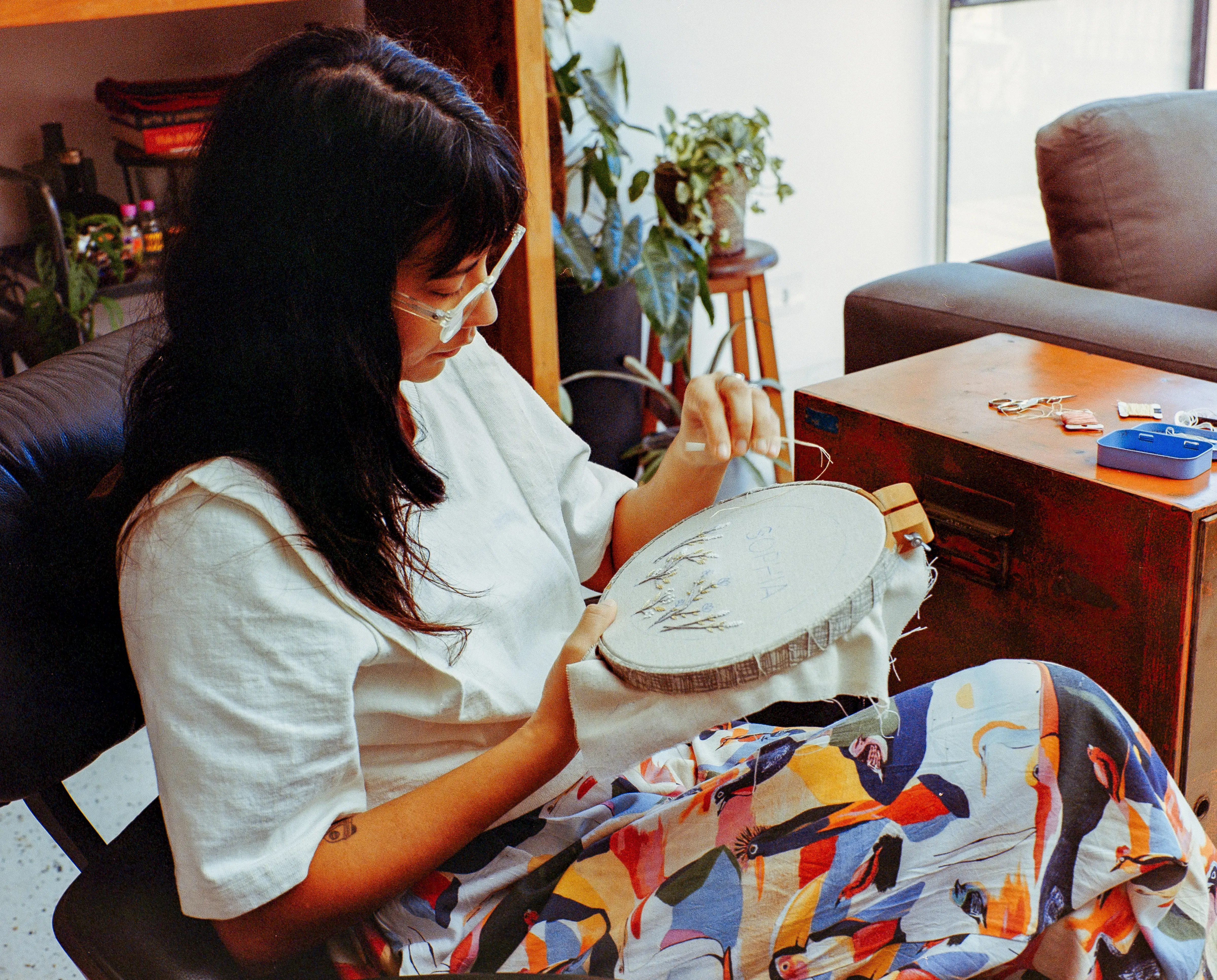 A woman with long dark hair and glasses sits in a black chair, focusing intently on embroidering a design inside a wooden hoop. She wears a white top and colorful patterned pants, with a wooden trunk and plants in the background.