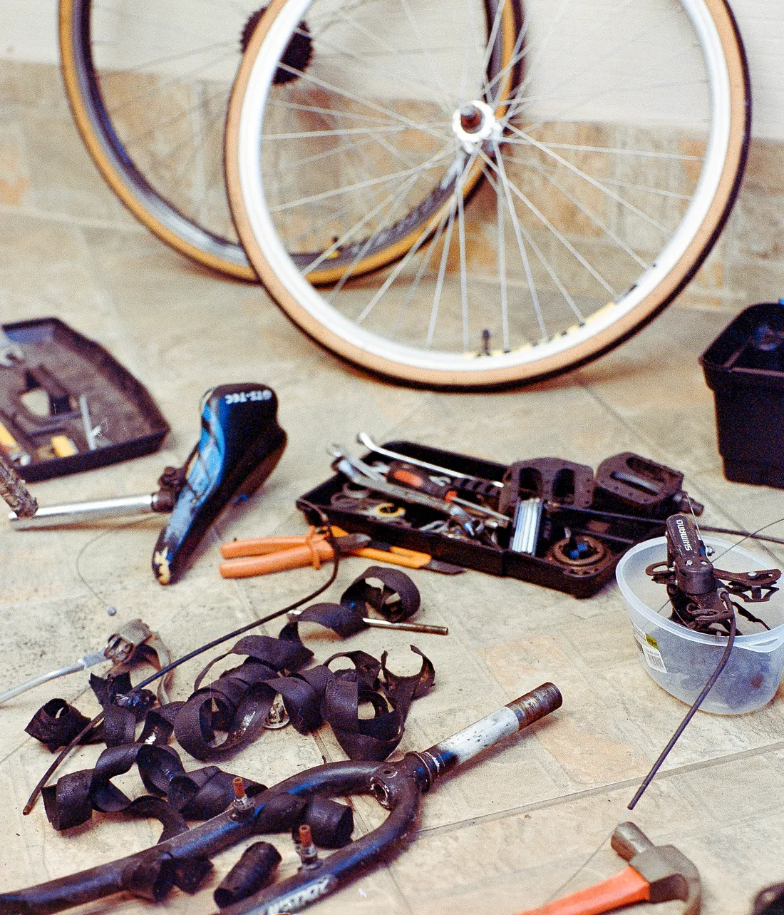 Disassembled bicycle parts and tools scattered on a tiled floor. Visible items include two wheels, a seat, a front fork, wrenches, pliers, a hammer, and small containers holding components.