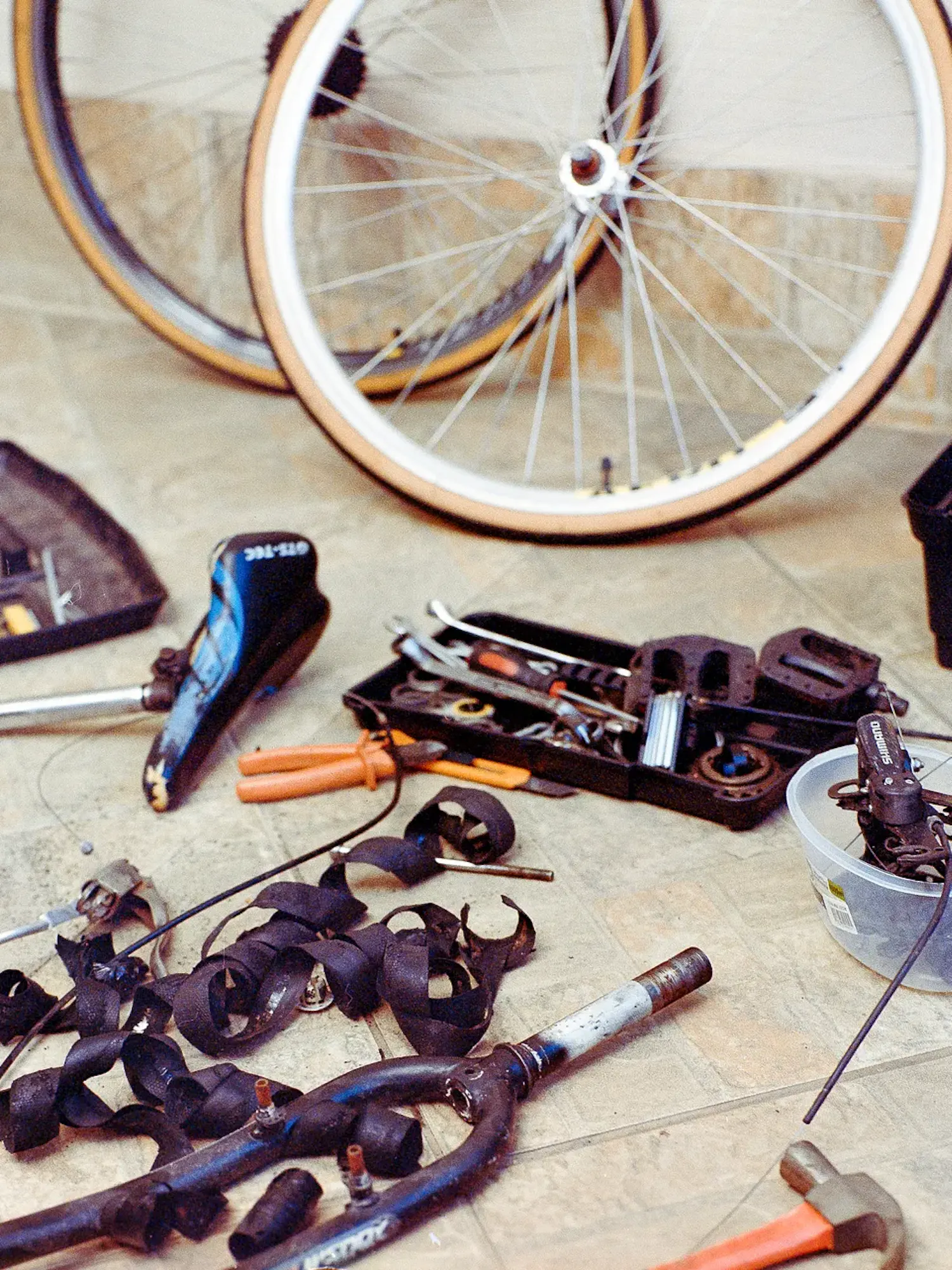 Disassembled bicycle parts and tools scattered on a tiled floor. Visible items include two wheels, a seat, a front fork, wrenches, pliers, a hammer, and small containers holding components.
