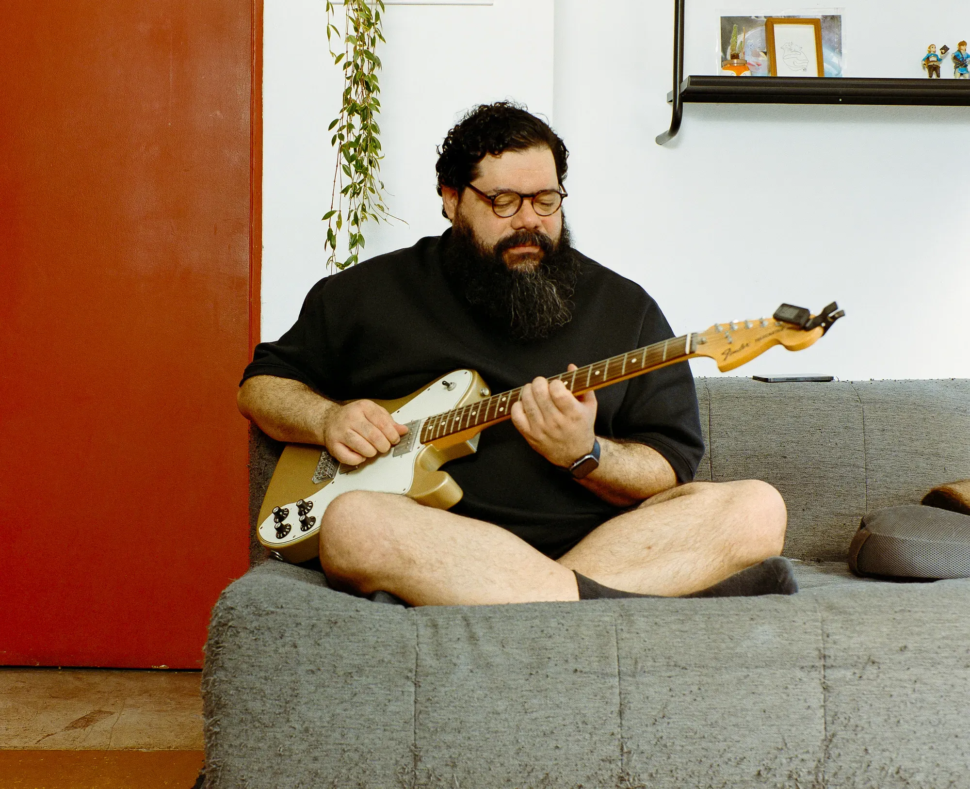 A man with a full beard, curly hair, and glasses sits cross-legged on a grey sofa, playing a gold Fender Telecaster electric guitar. A vibrant red door and a trailing green plant are in the background.