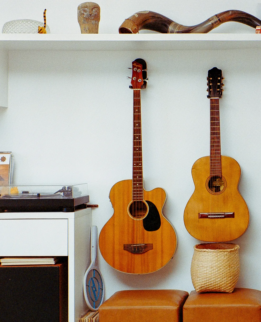 An acoustic bass and a guitar hang on a white wall next to a turntable on a cabinet. A shelf above holds decorative objects like a large horn, while leather stools and a woven basket rest below.