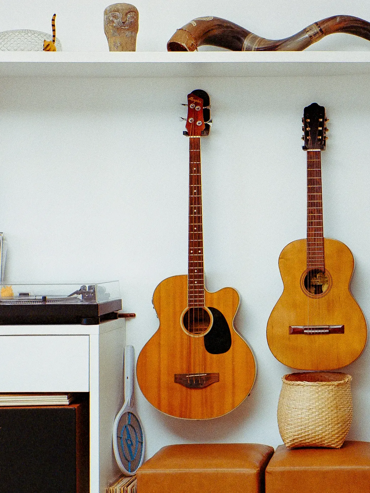 An acoustic bass and a guitar hang on a white wall next to a turntable on a cabinet. A shelf above holds decorative objects like a large horn, while leather stools and a woven basket rest below.