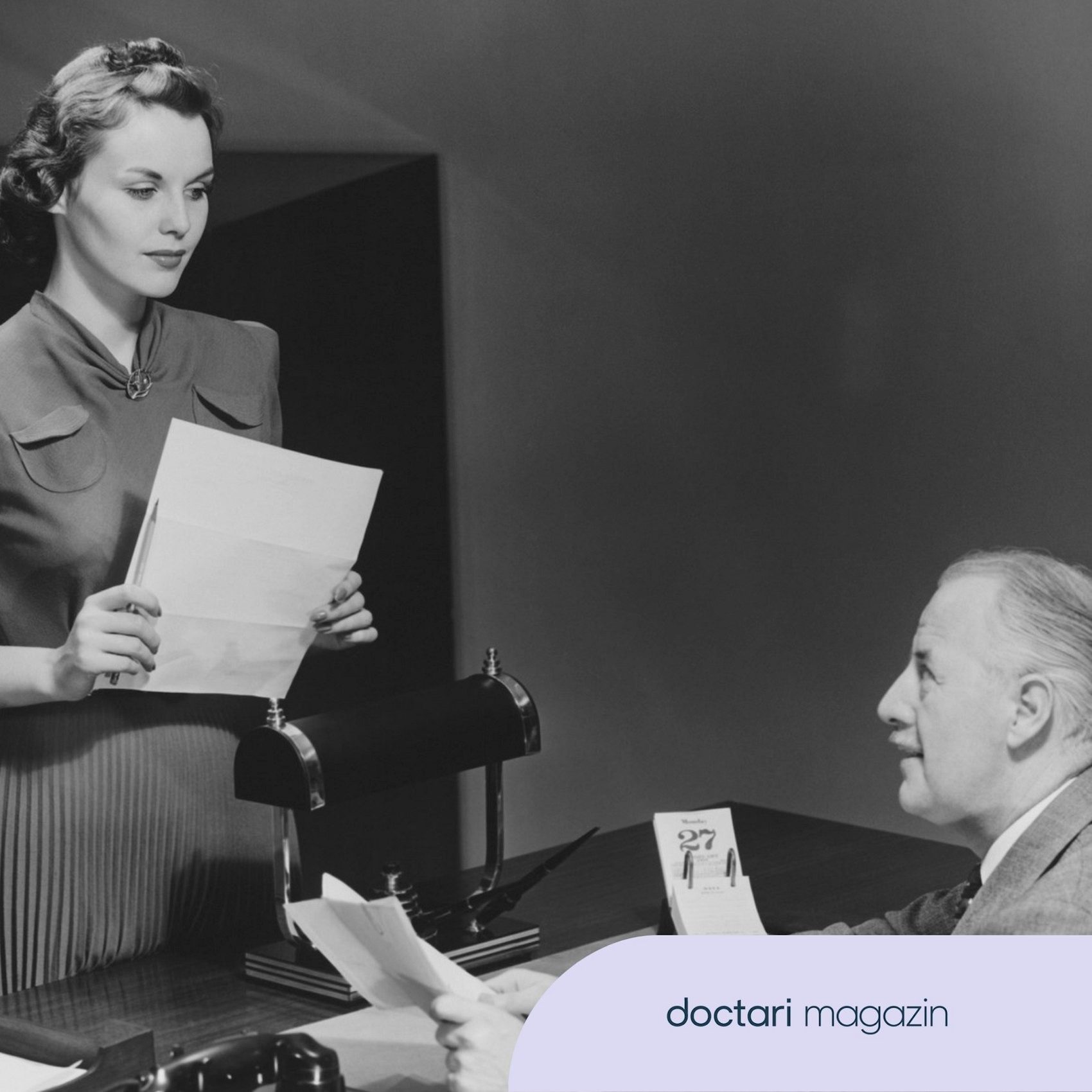 Eine Sekretärin aus den Fünfzigerjahren steht vor dem Schreibtisch ihres Chefs.