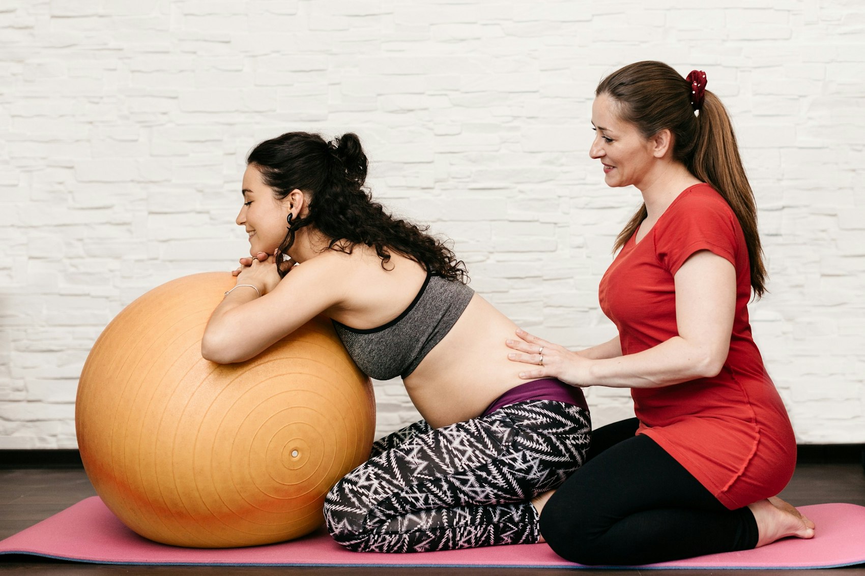 Eine Hebamme massiert einer Schwangeren den Rücken, während diese sich über einen Gymnastikball lehnt.