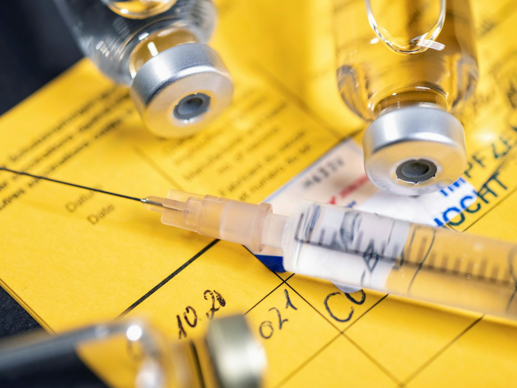 Eine Spritze sowie zwei Ampullen liegen auf einem gelben Impfpass.