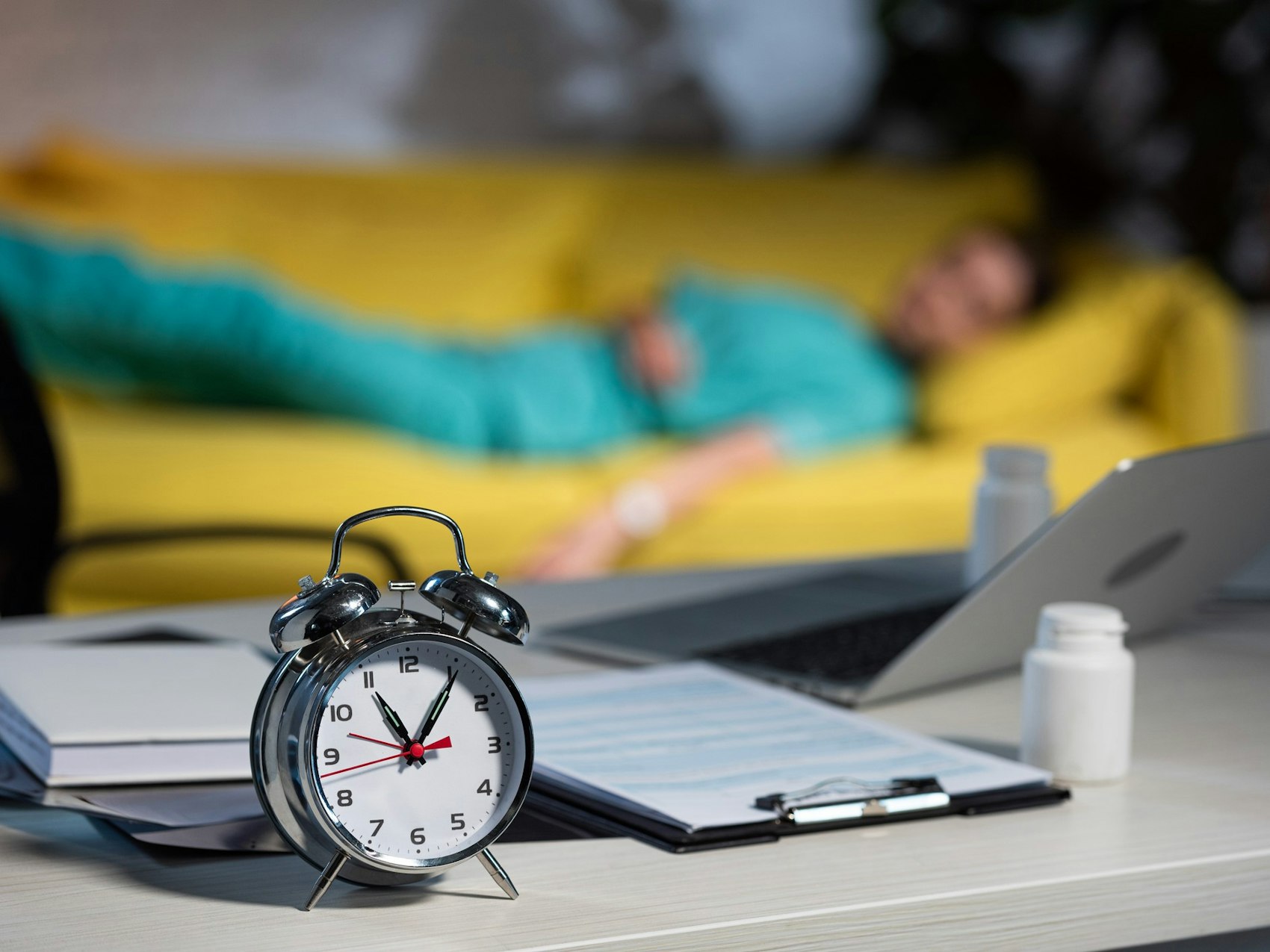 Ein Wecker steht auf einem Schreibtisch, im Hintergrund schläft eine Ärztin