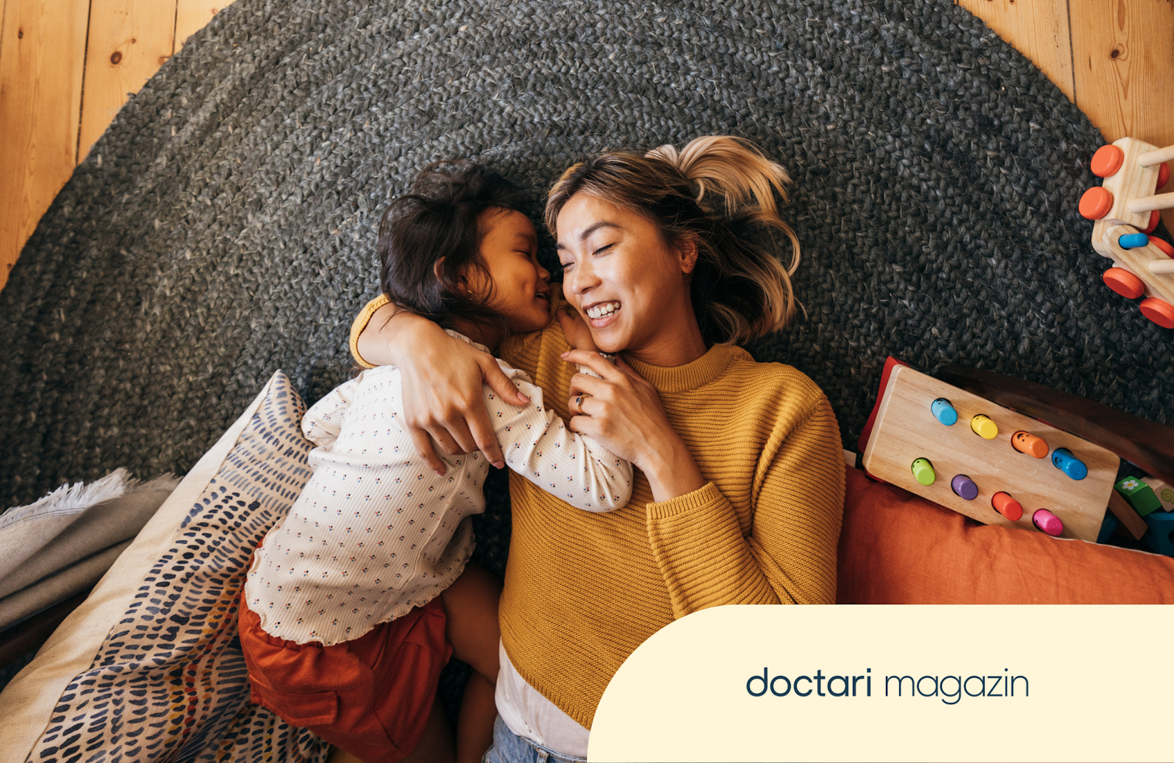 Eine junge Mutter liegt zusammen mit ihrer Tochter auf dem Boden. Sie halten sich im Arm und lachen