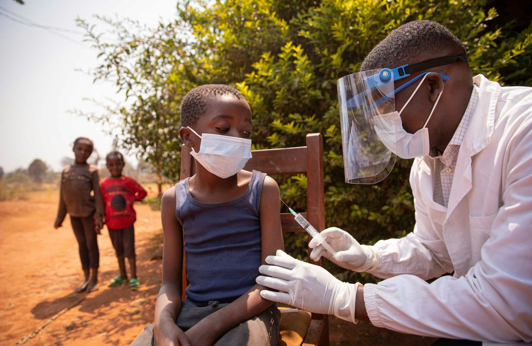 Arzt mit Schutzvisier impft ein Kind in einem afrikanischem Dorf