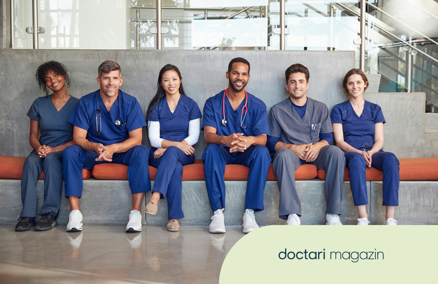 Mehrere Pflegekräfte im Kasack sitzen nebeneinander auf einer Bank in einem Krankenhausflur.