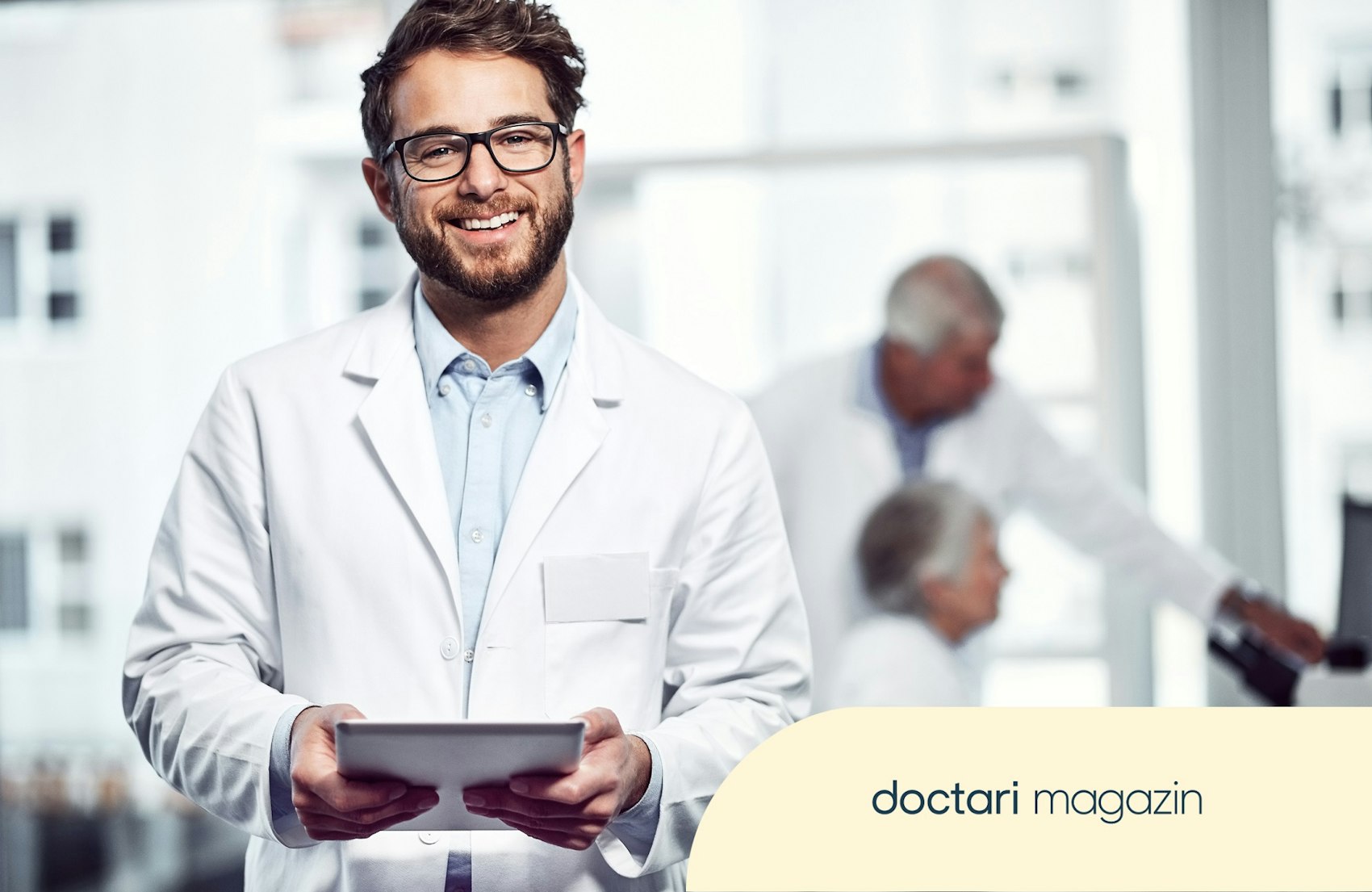 Junger Arzt mit Brille hält ein Tablet in der Hand und blickt freundlich in die Kamera.