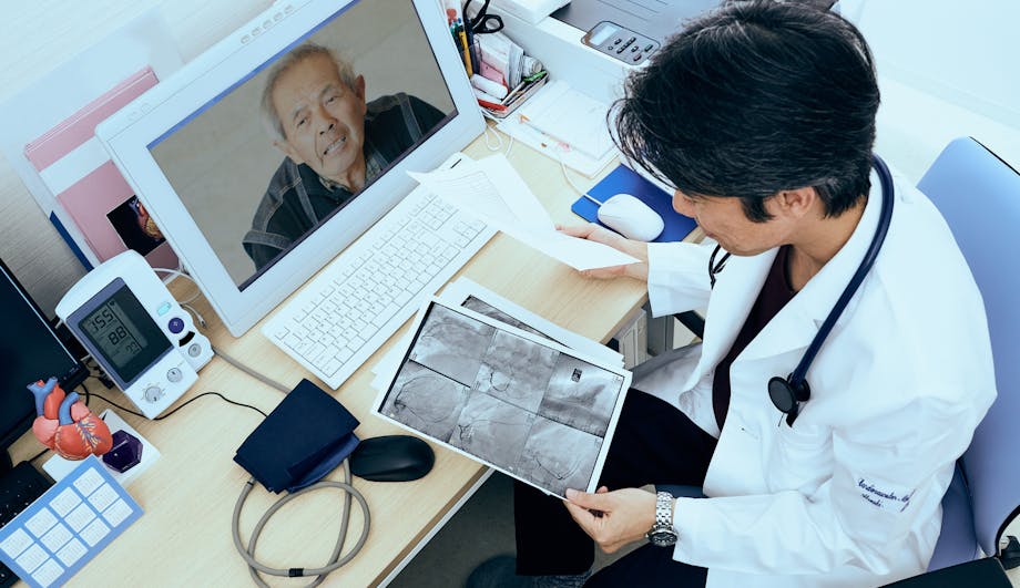 View on a doctors desk going through files while he is on a telemedicine call with his patient on a desktop computer