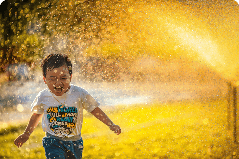 Atmosphärisches Kind unter dem Regen eines Wassersprinklers, was durch die Sonne gold gefärbt ist