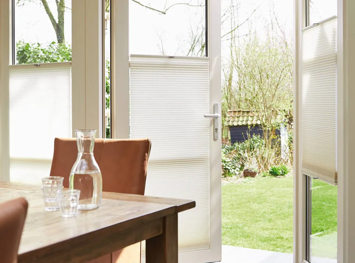 Cream Duette® honeycomb blinds on patio doors in a bright dining room overlooking a garden.