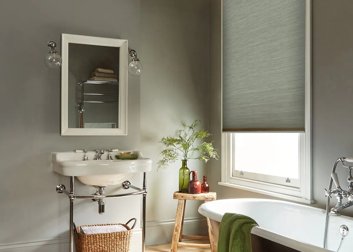 Light grey Duette honeycomb blind on a bathroom window beside a sink, mirror and freestanding bath.