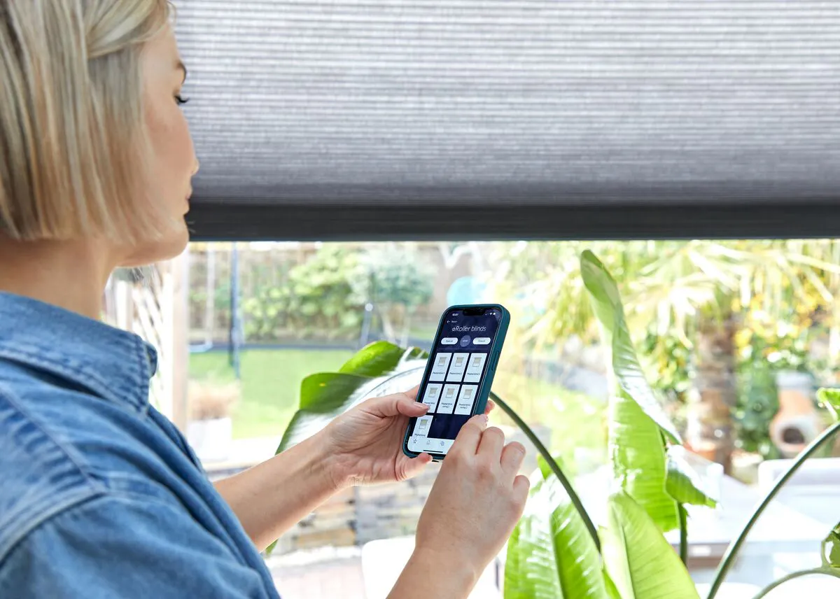 Person adjusting grey Duette honeycomb blinds using a smartphone in a bright room with plants.