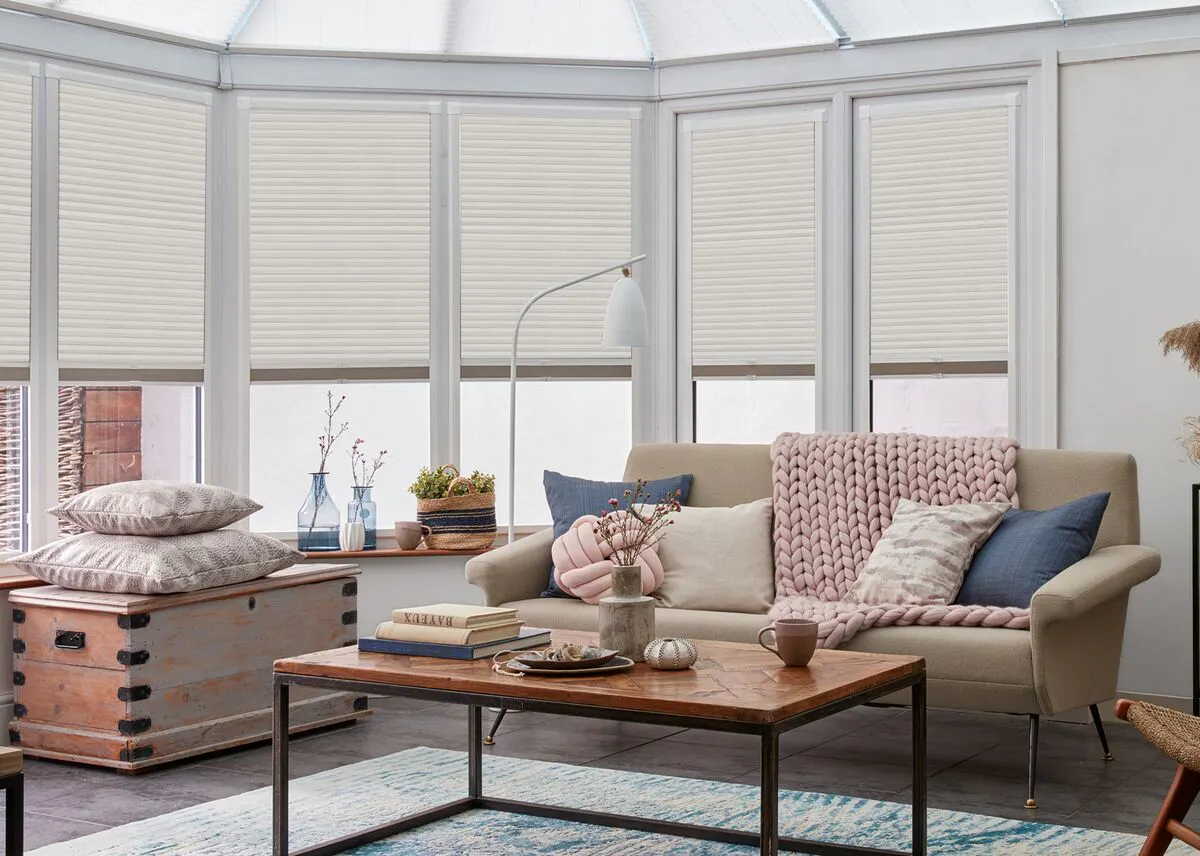 Beige Duette honeycomb blinds on conservatory windows surrounding a bright seating area with plants and a corner sofa.