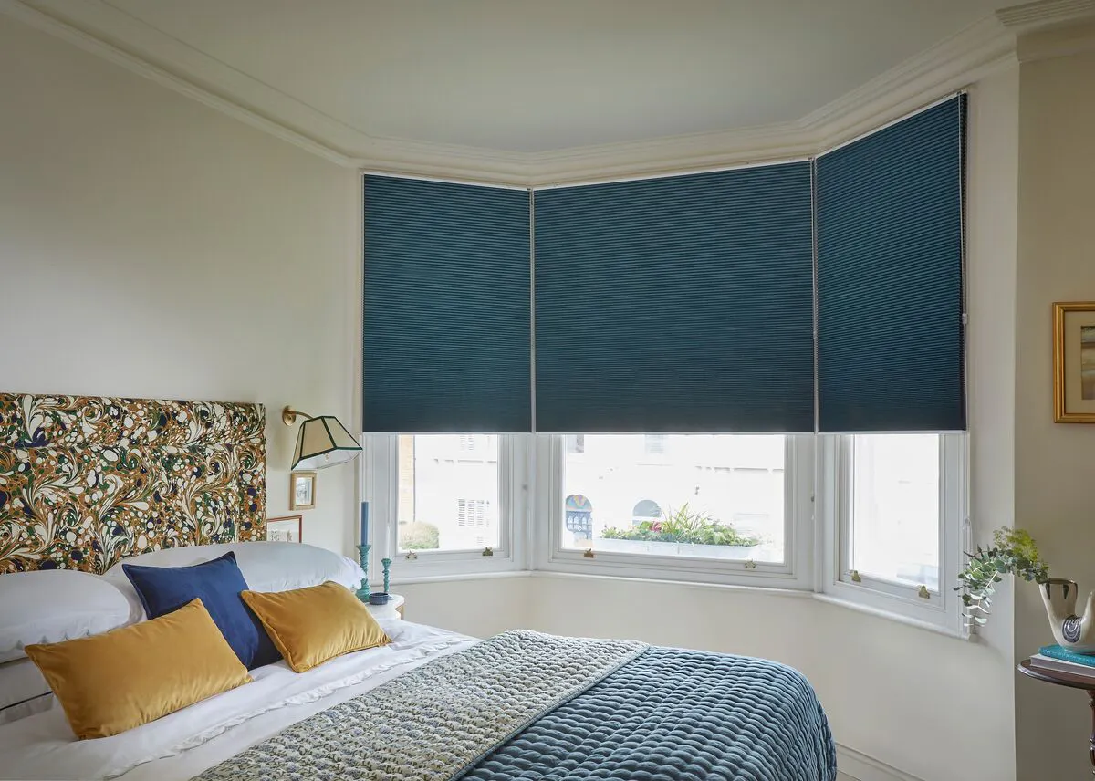 Deep blue Duette honeycomb blinds on bay windows in a stylish bedroom with patterned bedding and a decorative headboard.