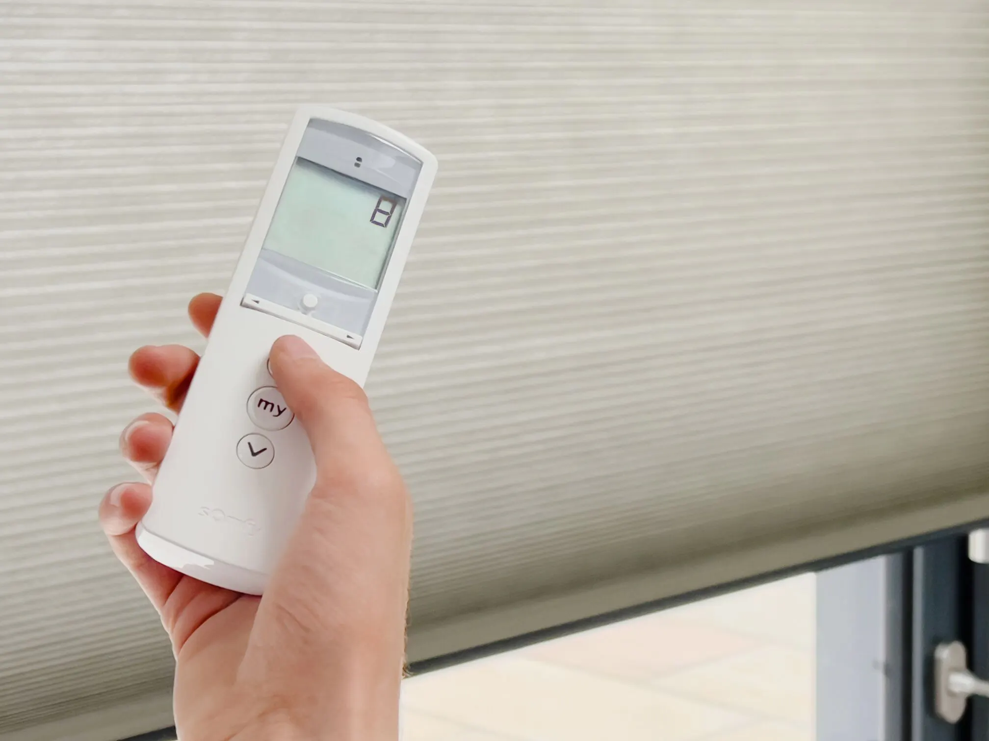 Hand holding a remote control in front of a light grey Duette honeycomb blind.