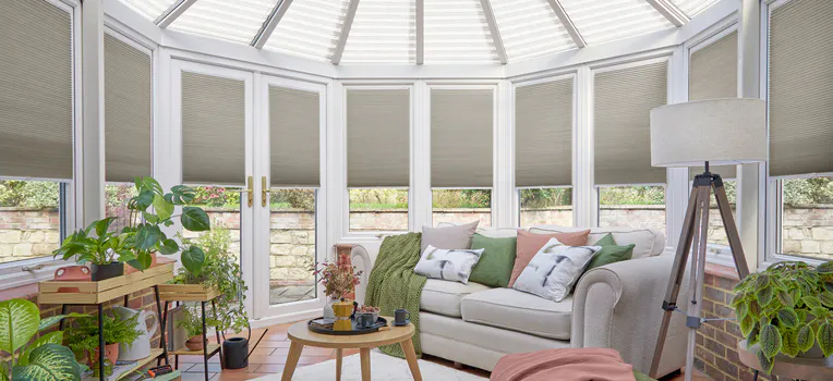 Beige Duette honeycomb blinds on conservatory windows surrounding a bright seating area with plants and a neutral sofa.