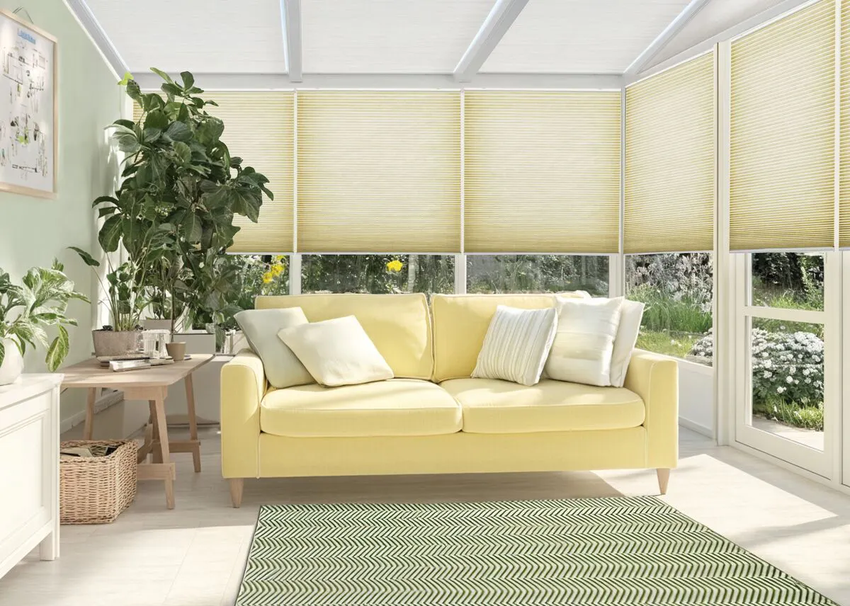 Soft yellow Duette honeycomb blinds on conservatory windows behind a light yellow sofa and indoor plants.