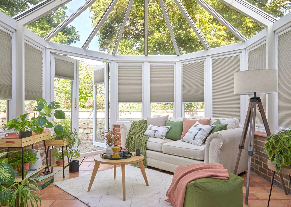 Beige Duette honeycomb blinds on conservatory windows around a bright seating area with plants and a cosy sofa.