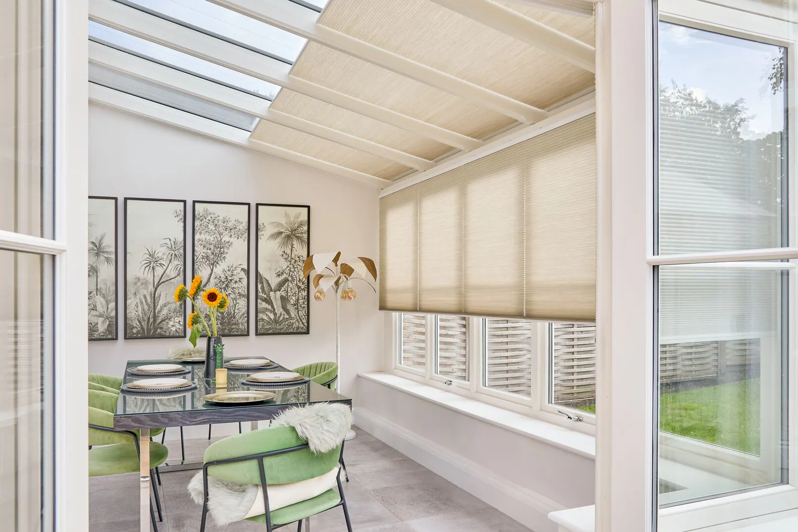 Beige Duette honeycomb blinds on conservatory windows above a dining table with green chairs and botanical artwork.