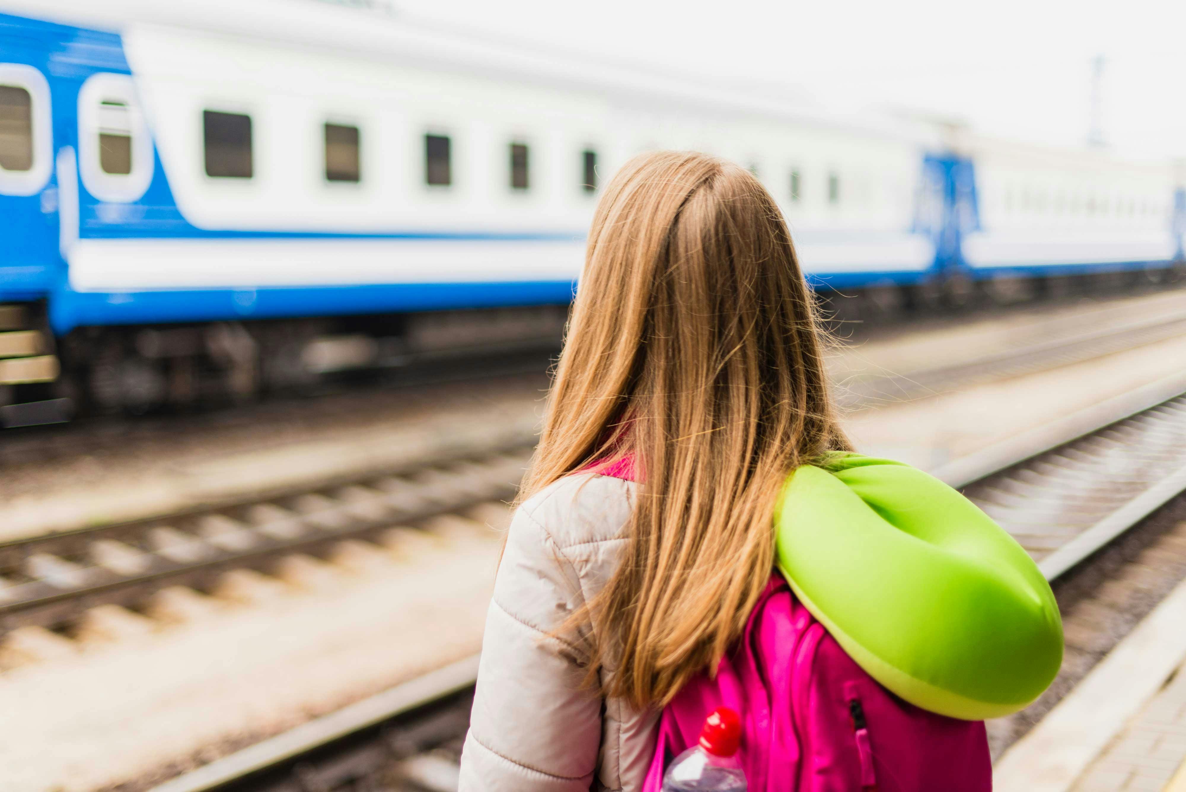  una niña espera el tren con una almohada de viaje y una mochila