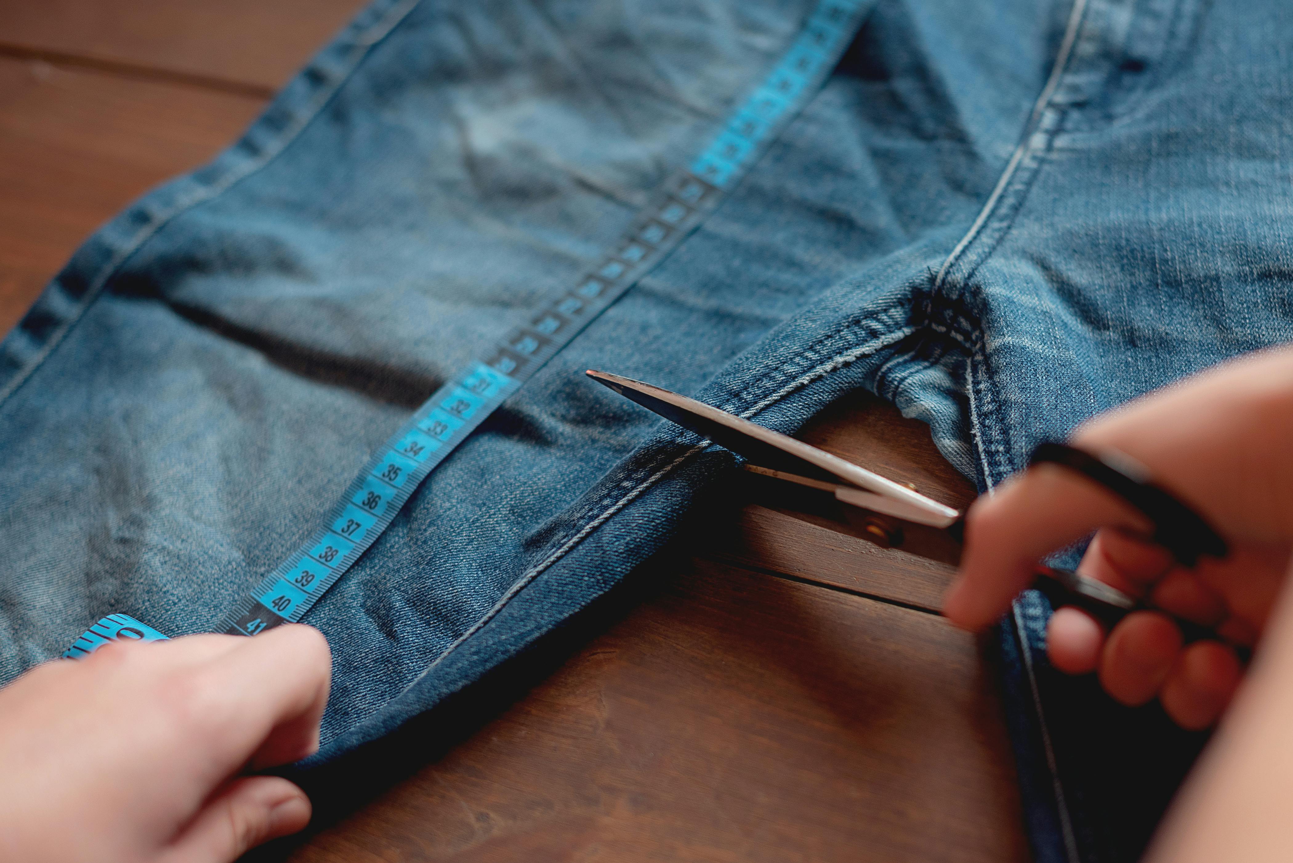 hands cutting denim jeans into shorts using a measuring tape and scissors