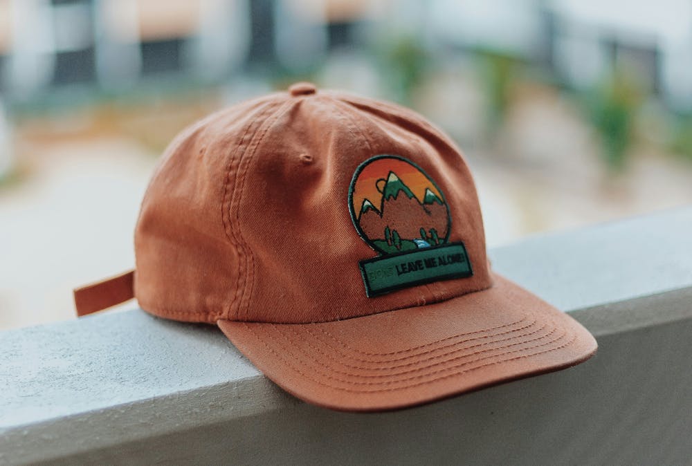 A brown baseball cap with an embroidered patch laying on a white railing A brown baseball cap with an embroidered patch laying on a white railing