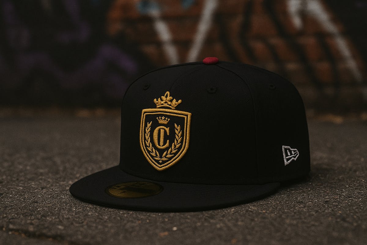 Black fitted cap with a gold embroidered crest and red top button, photographed on a street with a graffiti-covered wall in the background Black fitted cap with a gold embroidered crest and red top button, photographed on a street with a graffiti-covered wall in the background