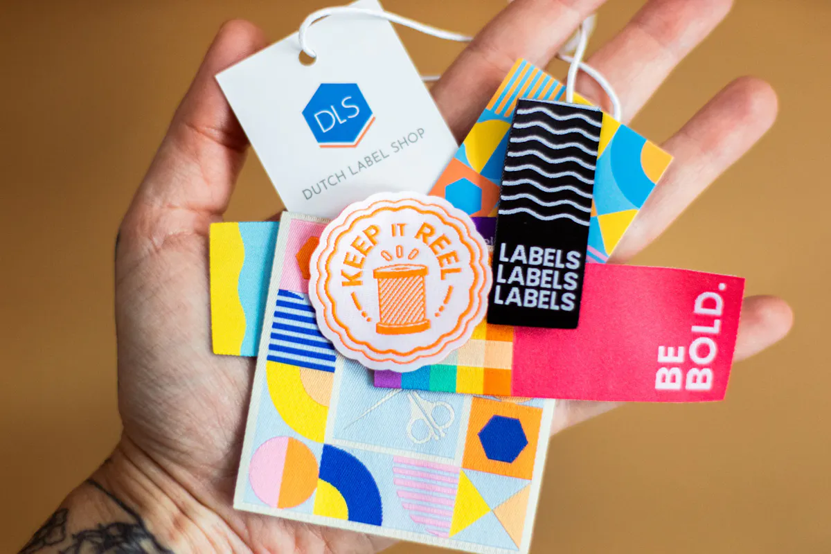  A hand holding a collection of colorful custom woven labels, fabric tags, and patches with bold geometric patterns and embroidered branding.