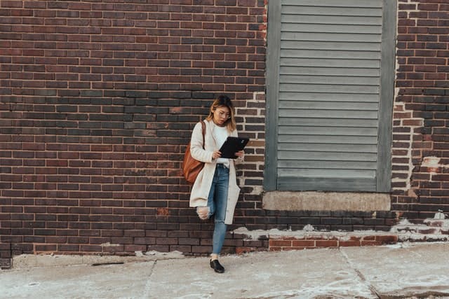 Woman on the sidewalk looking at a tablet leaning against a brick wall