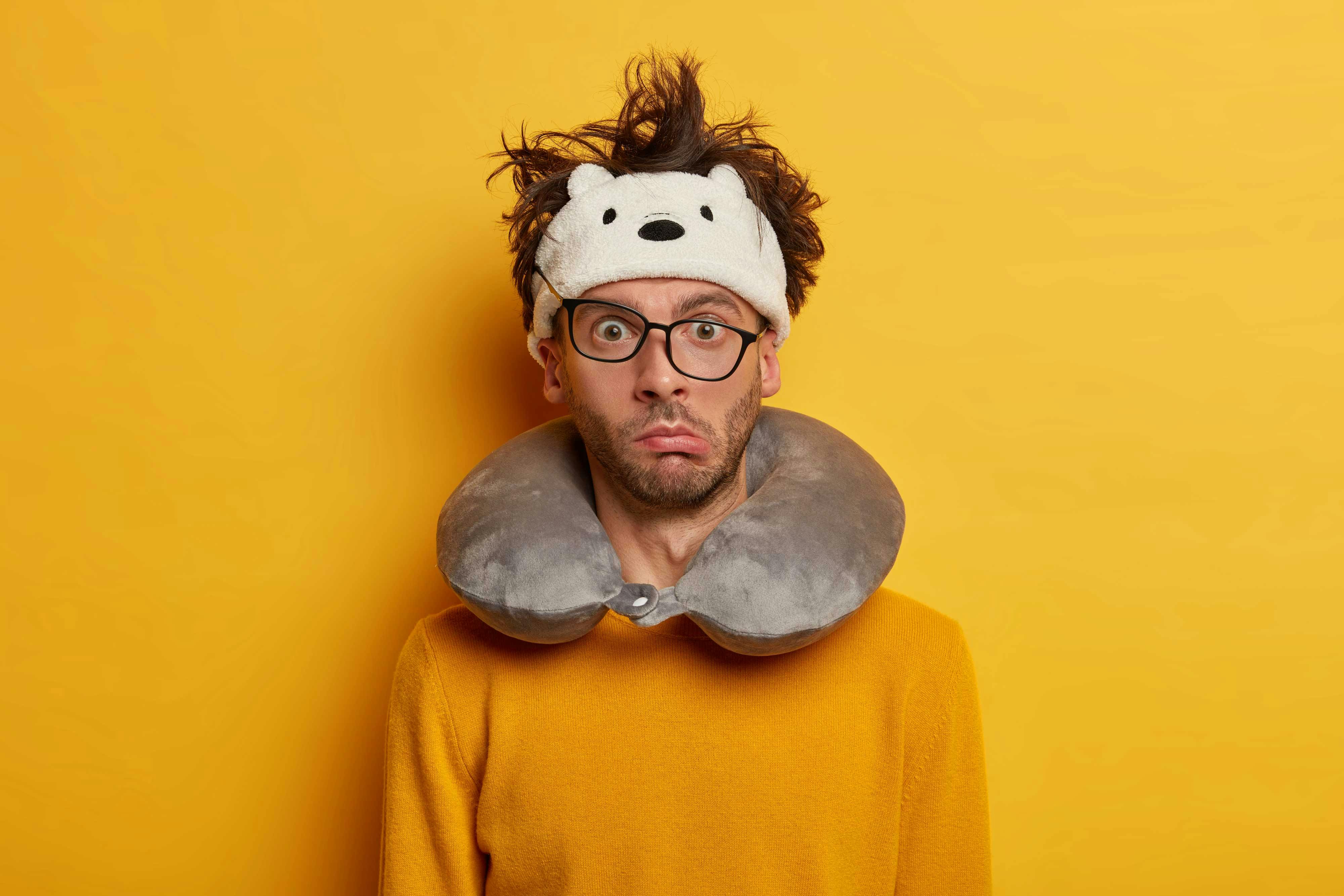 man wearing neck pillow with glasses askew and funny expression against bright yellow wall