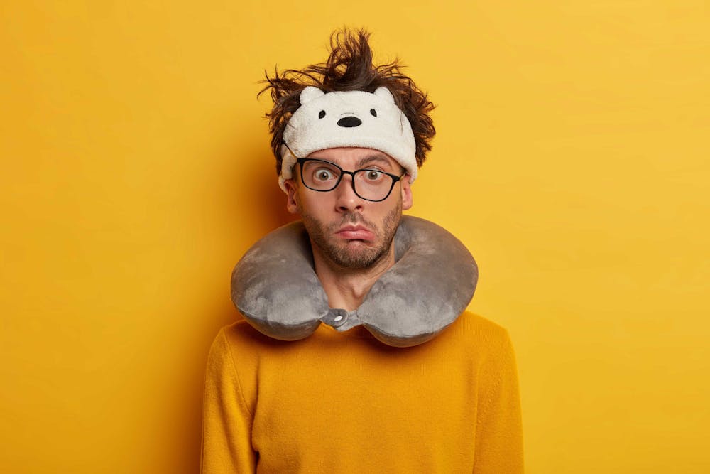 homme portant un coussin de voyage avec des lunettes de travers et une expression rigolote adossé contre un mur jaune vif homme portant un coussin de voyage avec des lunettes de travers et une expression rigolote adossé contre un mur jaune vif