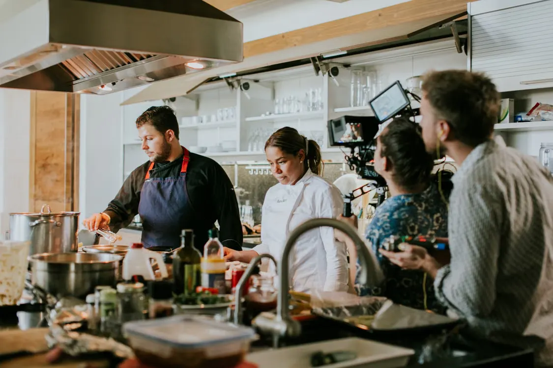 Restaurant chefs preparing food in a professional kitchen while a camera crew films the cooking process.