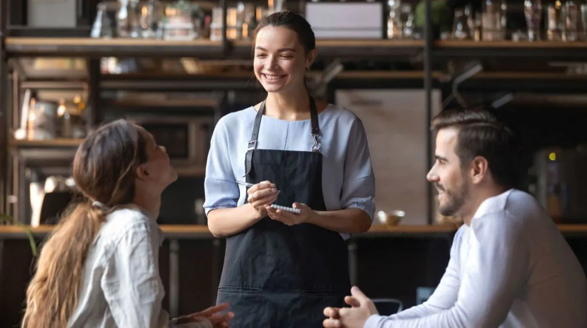 Smiling restaurant server taking an order and interacting with two guests seated at a restaurant table.