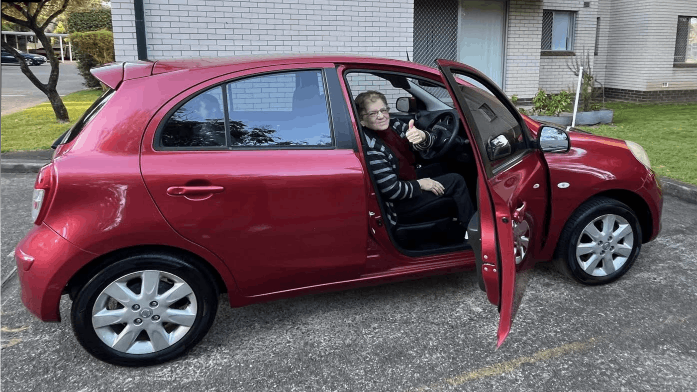 An older woman sitting in a plum-coloured Nissan Micra looking at the camera with her thumbs up