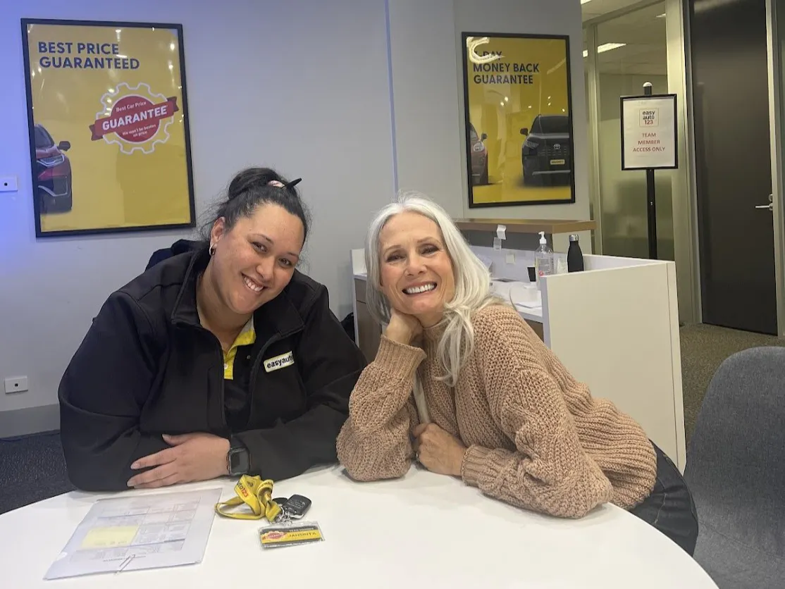 A customer and salesperson in a used car dealership are seated at a table and smiling happily