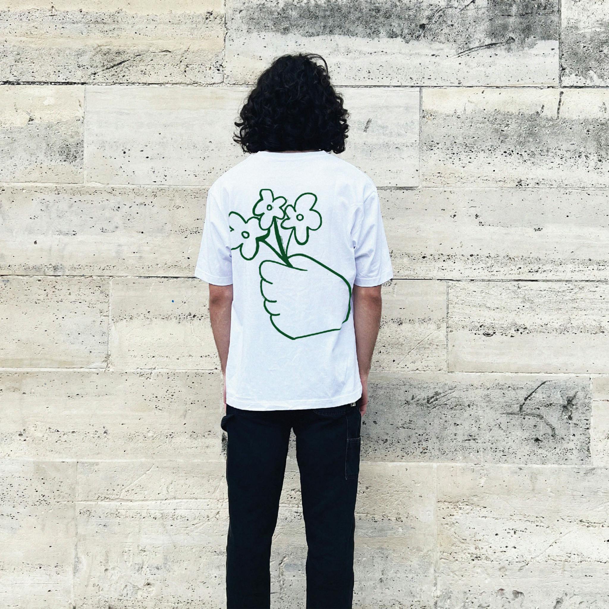 A man with long dark hair wearing a white T-shirt with a green outline drawing of a hand holding three flowers on the back.